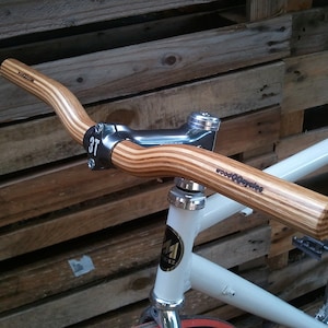 Può includere: Una bicicletta bianca con un manubrio in legno che dice "Wood2Cycles". Il manubrio è fatto di più strati di legno e ha uno stelo in metallo argentato.