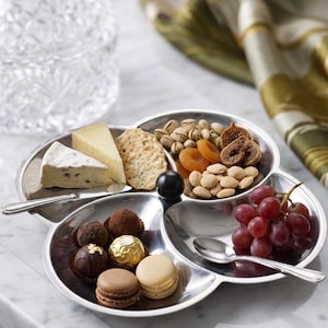 Peut inclure: Un plateau de service en métal argenté à trois compartiments contenant un assortiment de nourriture. Une section contient des morceaux de fromage et des biscuits, une autre des fruits secs et des noix, et la troisième des truffes et macarons.