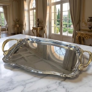 May include: A silver serving tray with gold handles, displayed on a marble surface. The tray has a decorative edge and curved handles. The tray is rectangular in shape and is suitable for serving food or drinks.