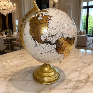 May include: A decorative globe featuring a white sphere with metallic gold continents and black text labels. The globe is supported by a gold-colored metal meridian ring and a circular pedestal base, displaying geographic details of the Americas.