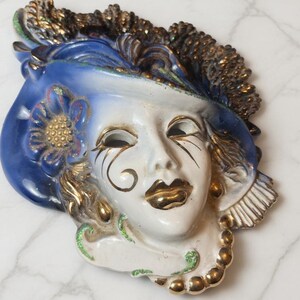 Peut inclure: Un masque en céramique avec un chapeau bleu et un visage blanc. Le masque a des accents dorés sur les lèvres, autour des yeux et sur la fleur. Le masque est orné d'un collier de perles dorées et d'accents verts.