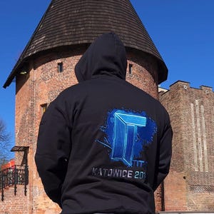 Puede incluir: Una sudadera negra con capucha que presenta un logotipo gráfico azul en la parte posterior con el texto "TITAN" y "KATOWICE 2019" impreso debajo. La sudadera se combina con pantalones vaqueros grises, mostrando un diseño llamativo.