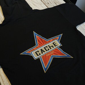 May include: A black hooded sweatshirt featuring a large embroidered star graphic on the back. The star is red with blue outlines and contains a gold rectangular banner with the black text CACHE and the smaller text CS GO printed below it.