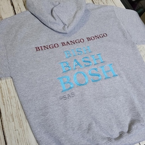 Puede incluir: Una sudadera con capucha gris con las frases bordadas "BINGO BANGO BONGO" en granate y "BISH BASH BOSH" en azul. La sudadera tiene una capucha con cordón y mangas cortas. Las letras "SAS" están bordadas debajo del texto azul.