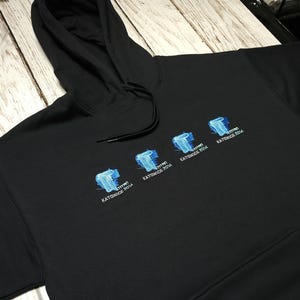 Puede incluir: Una sudadera negra con capucha que presenta tres logotipos bordados idénticos en el pecho. Cada logotipo muestra una letra T azul con el texto TITAN KATOWICE 2014 impreso debajo. La prenda incluye una capucha con cordón y un bolsillo frontal.