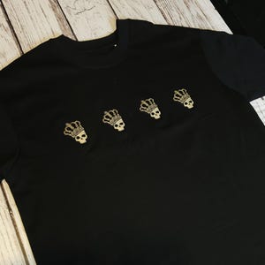 May include: A black short-sleeved t-shirt featuring four embroidered gold-coloured skull icons, each wearing a crown. The skulls are arranged in a horizontal row across the upper chest area of the garment, showcasing detailed stitching and metallic thread.