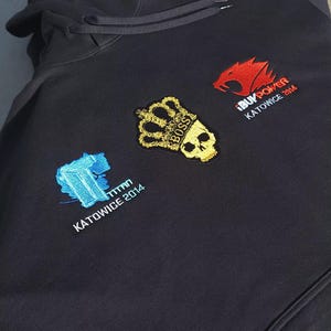 Könnte beinhalten: Schwarzer Kapuzenpullover mit gestickten Aufnähern. Aufnäher zeigen ein blau-weißes geometrisches Design, eine goldene Krone und einen Totenkopf sowie ein rotes Wolfslogo. Auf den Aufnähern steht "TITAN KATOWICE 2014", "BOSS" und "iBUYPOWER KATOWICE 2014".