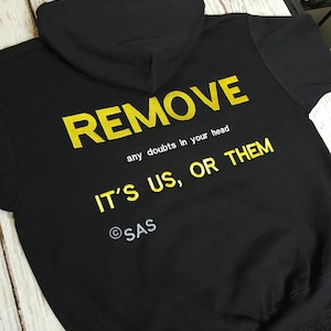 Puede incluir: Sudadera con capucha negra con texto amarillo. El texto dice "REMOVE" encima de "any doubts in your head" y "IT'S US, OR THEM". La sudadera también tiene el texto "©SAS".