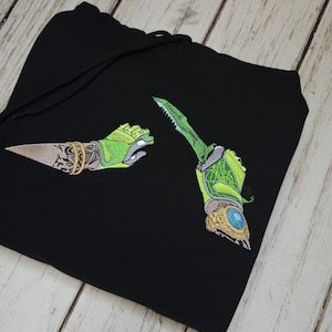 May include: A black hooded sweatshirt featuring an embroidered design of two arms wearing bright green gloves. One arm holds a sharp blade, while the other displays a detailed tattoo and a gold watch. The embroidery includes intricate patterns.