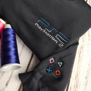 May include: Dark grey sweatshirt featuring a PlayStation 2 logo embroidered in blue and white. The left sleeve has embroidered controller symbols in various colours. The garment is on a wooden surface.