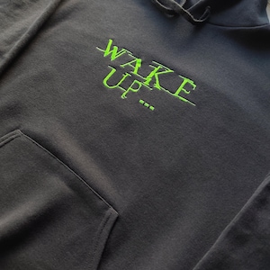 May include: A dark grey hoodie with a front pocket and a drawstring hood. The neon green embroidered text "WAKE UP..." is across the chest. The hoodie is made of a soft, comfortable material.