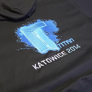 Puede incluir: Sudadera con capucha negra con un diseño bordado en azul y blanco. El diseño incluye la palabra "TITAN" y "KATOWICE 2014". La sudadera tiene capucha y está hecha de un material suave.