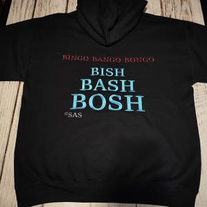 Puede incluir: Sudadera con capucha negra con las frases "BINGO BANGO BONGO" en rojo y "BISH BASH BOSH" en azul. La sudadera también tiene el logo "©SAS". La sudadera con capucha está hecha de un material suave.