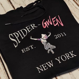 以下が含まれることがあります： 黒色のフード付きスウェットシャツ。刺繍のデザインは、白とピンクの「SPIDER-GWEN」の文字と、その下に「EST. 2011 NEW YORK」があります。スパイダーグウェンの漫画の画像も前面に刺繍されています。