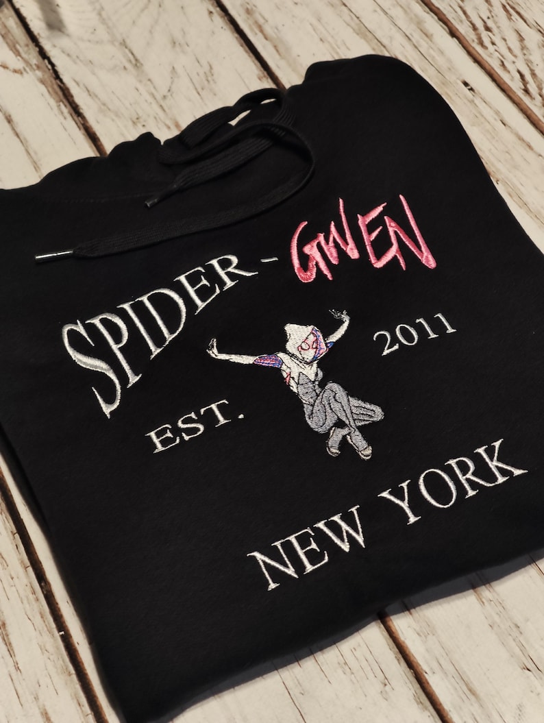 以下が含まれることがあります： 黒色のフード付きスウェットシャツ。刺繍のデザインは、白とピンクの「SPIDER-GWEN」の文字と、その下に「EST. 2011 NEW YORK」があります。スパイダーグウェンの漫画の画像も前面に刺繍されています。