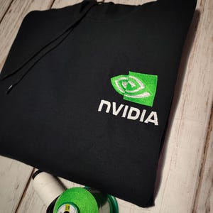 Puede incluir: Sudadera con capucha negra con el logotipo de NVIDIA bordado en blanco y verde. El logotipo presenta un diseño de ojo estilizado. La sudadera con capucha tiene un cordón y está hecha de un material suave. La imagen también muestra carretes de hilo.