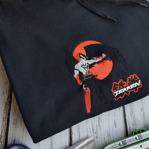 Może przedstawiać: Czarna bluza z kapturem z haftowanym wzorem przedstawiającym postać w akcji na pomarańczowym tle. Tekst "TEKKEN" jest wyświetlany w kolorze białym i czerwonym. Bluza ma sznurek i przednią kieszeń.