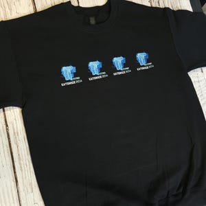 Puede incluir: Sudadera negra de cuello redondo con un diseño gráfico azul y blanco. El diseño presenta estructuras azules estilizadas con el texto "KATOWICE 2014" debajo. La sudadera está hecha de un material suave.