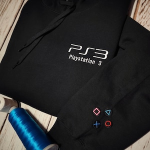 May include: Black hoodie with white embroidered "PS3 Playstation 3" text on the chest. The sleeve features embroidered pink, blue, green, and red controller symbols. A spool of blue thread is visible.