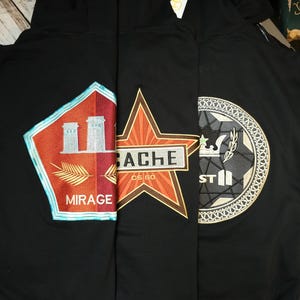 Op de afbeelding: Drie zwarte hoodies met geborduurde ontwerpen. De linker hoodie toont een vijfhoekige patch met de tekst MIRAGE. De middelste hoodie heeft een stervormige patch met de tekst CACHE CS GO. De rechter hoodie toont een cirkelvormig embleem.