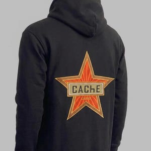 May include: A black hooded sweatshirt featuring a large embroidered design on the back. The design consists of an orange and gold star with a rectangular patch across the center displaying the text CACHE and the smaller text CS 60 below it.