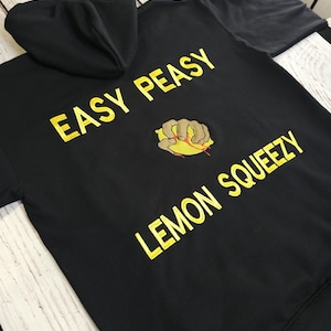Puede incluir: Sudadera con capucha negra con el texto amarillo "EASY PEASY LEMON SQUEEZY". La sudadera presenta un gráfico bordado de una mano exprimiendo un limón. Tiene una capucha con cordón y un bolsillo delantero.