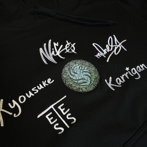 Könnte beinhalten: Ein schwarzer Kapuzenpullover mit weiß gestickten Signaturen und Texten, darunter "Niko", "Karrigan", "Yousuke" und "EESS". Ein runder Aufnäher mit einem grün-blauen geometrischen Design ist mittig zwischen den weißen Stickereien platziert.