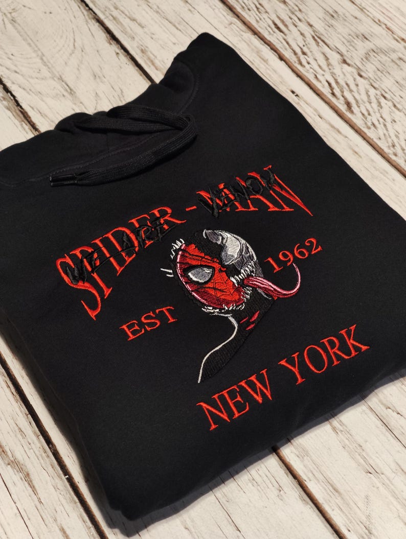 以下が含まれることがあります： 赤色の刺繍テキストとグラフィックが施された黒いフード付きスウェットシャツ。「SPIDER-MAN」が、ヴェノムと融合したスパイダーマンの画像の上にあり、「EST 1962 NEW YORK」が下にあります。フードにはドローコードが付いています。