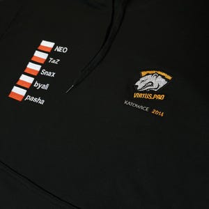May include: A black hooded sweatshirt featuring an embroidered Virtus.pro logo with a bear graphic, the text "VIRTUS.PRO KATOWICE 2014", and a vertical list of five Polish flag icons next to the names NEO, TaZ, Snax, byali, and pasha in white thread.