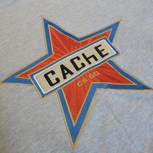 May include: A light gray fabric featuring an embroidered star design. The star has a bright orange center with radiating lines, outlined in blue and gold thread. A rectangular beige patch across the center displays the black text CACHE and CS GO.