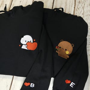 Könnte beinhalten: Zwei schwarze Kapuzenpullover mit gestickten Motiven. Ein Hoodie zeigt einen weißen Panda mit einem roten Herz, der andere einen braunen Bären mit einer Rose. Jeder Ärmel ist mit einem roten Herz und den Buchstaben B bzw. E bestickt.