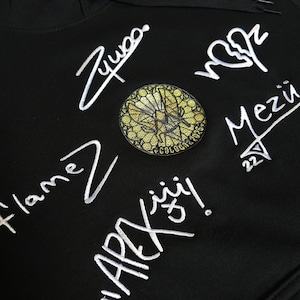 May include: A black hoodie featuring white embroidered signatures and text including "Zywoo", "Flamez", "APEX", and "Mezü 22". A circular patch with a gold and black geometric pattern is centered among the white embroidered designs.