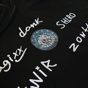 Könnte beinhalten: Ein schwarzer Kapuzenpullover mit weißem Sticktext, der "donk", "SHIRO", "twik", "zonk" und "gixx" lautet. In der Mitte befindet sich ein kreisförmiger Stickaufnäher mit einem blau-weißen Buntglasmuster um einen zentralen Tierkopf.