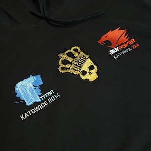 Puede incluir: Sudadera con capucha negra con parches bordados. Los parches incluyen una corona y una calavera doradas con el texto "BOSS", un logotipo rojo con el texto "iBUYPOWER KATOWICE 2014" y un diseño azul con el texto "TITAN KATOWICE 2014".