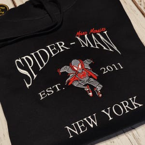 以下が含まれることがあります： 黒色のフード付きスウェットシャツ。白い刺繍で「SPIDER-MAN」の文字とスパイダーマンのグラフィックが描かれています。キャラクターの画像の下には「EST. 2011 NEW YORK」と書かれています。グラフィックは赤、グレー、黒です。