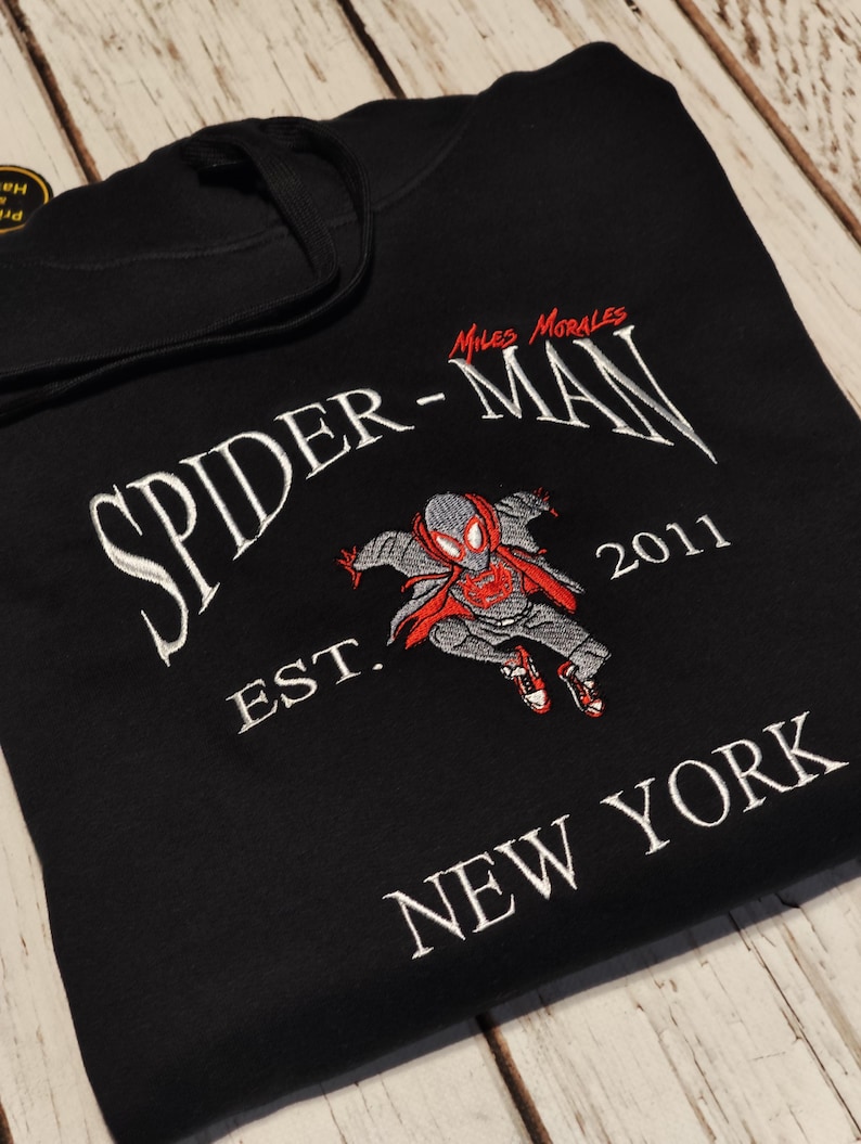 以下が含まれることがあります： 黒色のフード付きスウェットシャツ。白い刺繍で「SPIDER-MAN」の文字とスパイダーマンのグラフィックが描かれています。キャラクターの画像の下には「EST. 2011 NEW YORK」と書かれています。グラフィックは赤、グレー、黒です。