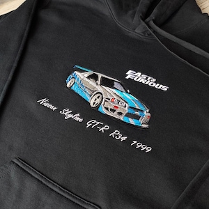 Peut inclure: Sweat à capuche noir avec un motif brodé d'une voiture Nissan Skyline GT-R R34 argent et bleu. Le texte "Fast & Furious" et "Nissan Skyline GT-R R34 1999" sont également brodés sur le sweat à capuche.