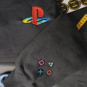 Peut inclure: Sweat-shirt gris foncé avec un logo PlayStation brodé en rouge, bleu, jaune et vert. La manche présente des symboles de boutons de manette brodés en rose, bleu, vert et orange. Vêtement sur le thème du jeu vidéo.