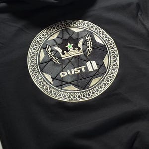 May include: A black hooded sweatshirt featuring a large circular embroidered patch on the back. The patch displays a crown, a small green star, and the text DUST in white lettering, surrounded by a geometric pattern and a decorative border.