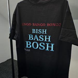 Puede incluir: Camiseta negra con las frases bordadas "BINGO BANGO BONGO" en rojo y "BISH BASH BOSH" en azul claro. La camiseta tiene cuello redondo y mangas cortas. La marca "SAS" está bordada en la parte inferior.
