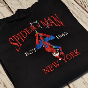 以下が含まれることがあります： 黒いフード付きスウェットシャツ。スパイダーマンのデザインが刺繍されています。「SPIDER-MAN NEW YORK」の文字が赤色で、1962年とサンタ帽をかぶったスパイダーマンが描かれています。スウェットシャツは木製の表面に折りたたまれています。