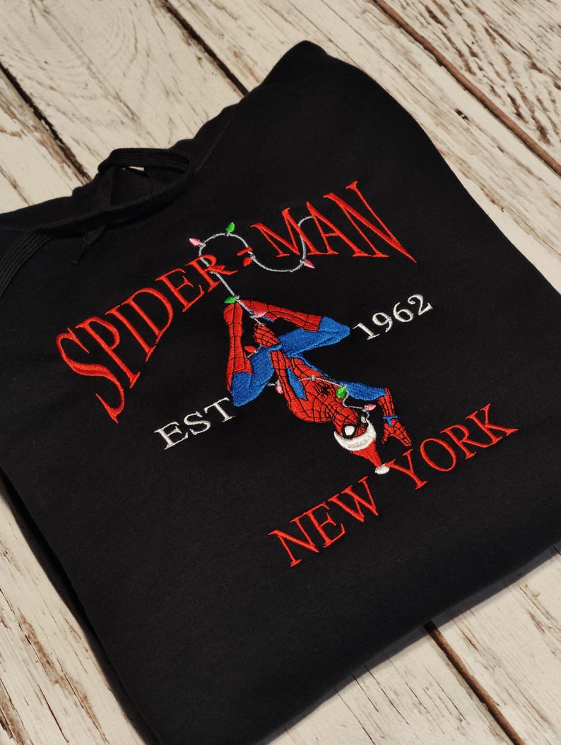 以下が含まれることがあります： 黒いフード付きスウェットシャツ。スパイダーマンのデザインが刺繍されています。「SPIDER-MAN NEW YORK」の文字が赤色で、1962年とサンタ帽をかぶったスパイダーマンが描かれています。スウェットシャツは木製の表面に折りたたまれています。