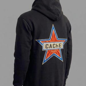 May include: A black hooded sweatshirt featuring a large embroidered star on the back. The star has orange and red sunburst rays, a blue border, and a central rectangular patch with the text CACHE and the smaller text CS GO below it.