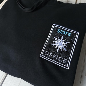Puede incluir: Una sudadera con capucha negra que presenta un parche rectangular bordado en el pecho. El parche tiene un fondo negro con un borde blanco, un gráfico de copo de nieve blanco en el centro, el número 52375 en azul claro y la palabra OFFICE en blanco.