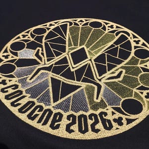 May include: Black fabric with a gold embroidered design. The design is a circular stained-glass style with a figure and the text "COLOGNE 2026". The embroidery includes silver and olive green accents.