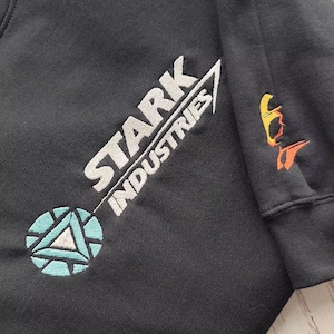 Könnte beinhalten: Schwarzes Sweatshirt mit dem Stark Industries Logo, gestickt in Weiß, Hellblau und Orange. Das Design beinhaltet den Stark Industries Text und ein kreisförmiges Emblem. Ein kleineres Emblem befindet sich am Ärmel.
