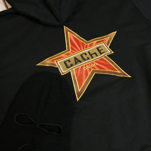 May include: Black hoodie with a star-shaped emblem featuring the word "CACHE" in a rectangular box, and the text "CS GO". A black balaclava is also visible.