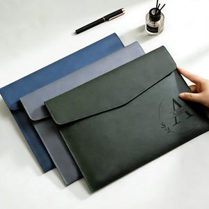 May include: Three leather document holders in blue, grey, and green. The green holder has the name "Albert" and the letter "A" printed on it. A black pen and a small bottle of fragrance are in the background.