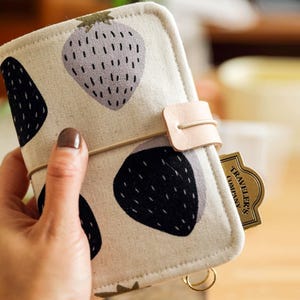 May include: A beige fabric journal cover with a repeating strawberry pattern in black and gray. The cover has a leather closure and a gold-colored tag that reads "Traveler's Company".