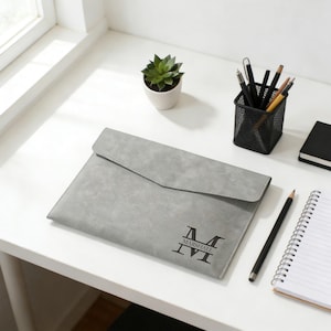 May include: A gray faux leather document holder with a flap closure. The holder features the name "MARSHALL" and the letter "M" in a stylized font. A small potted succulent, pens, pencils, and a notebook are also on the white desk.